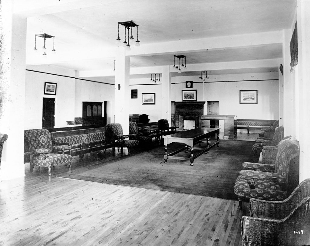 The interior lounge from a ward at Essondale Hospital. Benches and chairs line either side of the room. There is a table between the lines of chairs, and a fireplace at the back of the room. 
