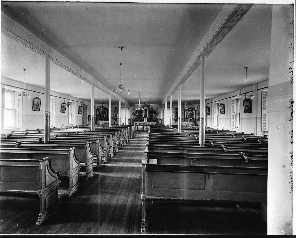 From the back of the room, the photograph captures the chapel at Longue Pointe Asylum in 1911. The long room was filled with rows of pews until the sanctuary. There were windows on either side of the room intermixed with holy paintings.