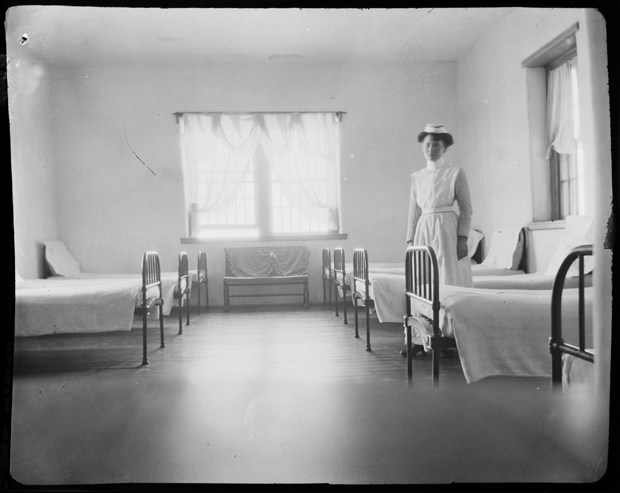 This image is of a smaller patient ward with eight beds shown. There is a bench in front of a window at the back of the room. A nurse stands between beds in a white cap and apron.