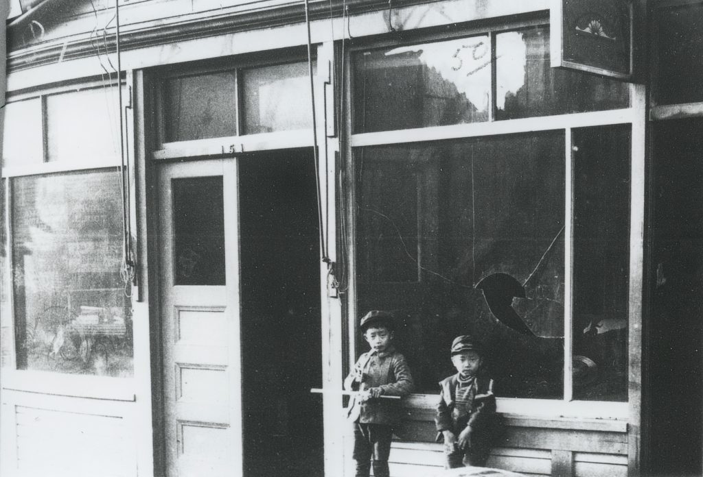 Two young boys are out front of a building that was damaged during the riot of 1907. The window behind them is broken and the door to the left is ajar.