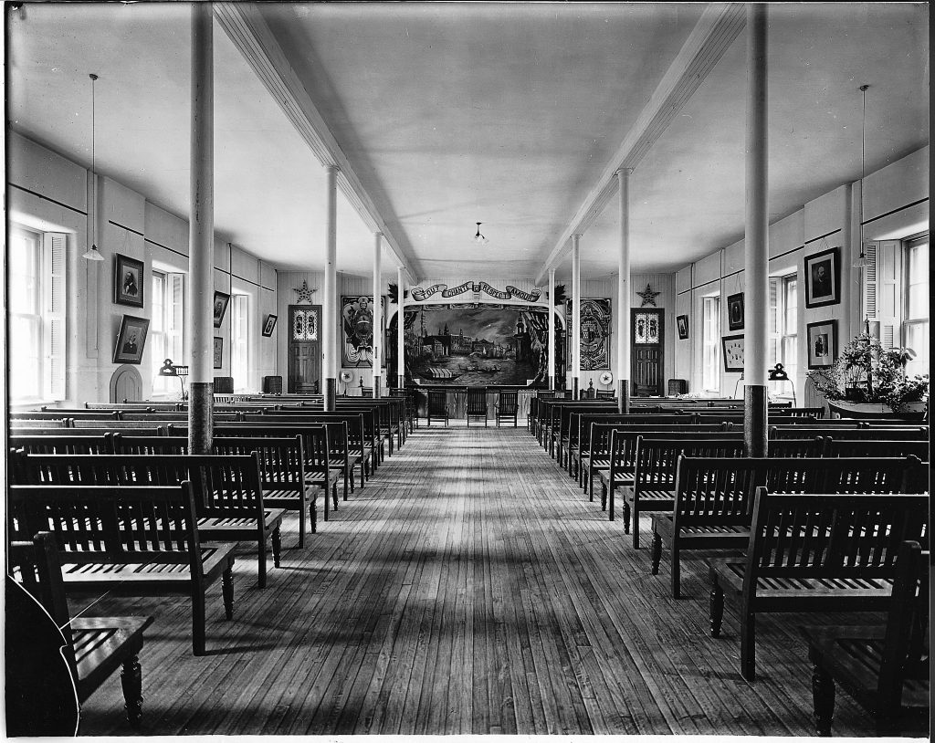 Captured from the back, wooden benches line the long room until the theater stage. There are windows on either side of the room intermixed with portraits. 