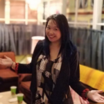 Smiling apparent young woman with long black hair and arms and hands outstreched standing in living room set. hair
