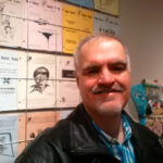 Head and shoulders shot of apparent man with mustache and goatee smiling with background of posters on wall behing.