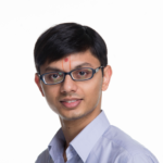 Head and shoulders of smiling young apparent man with short dark hair, glasses and red dot on forehead looking into the camera.
